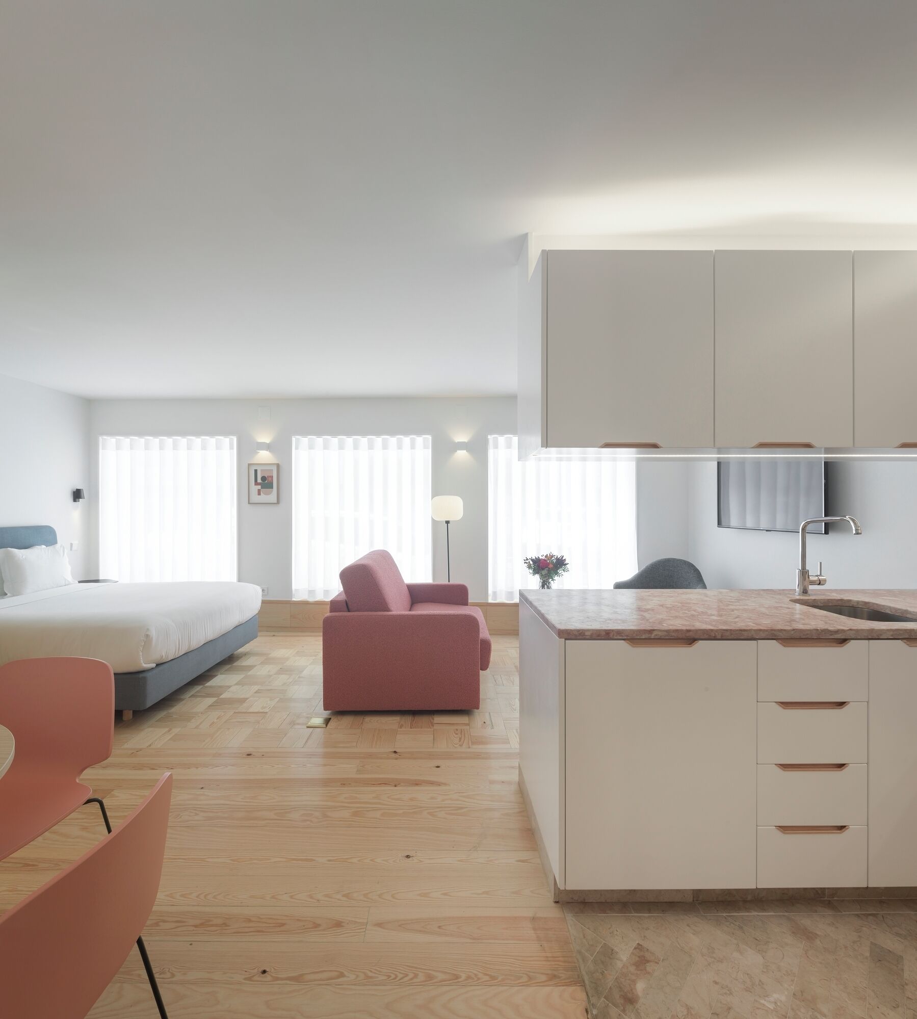 A modern hotel room with a bed, a pink armchair, a small kitchen area, and a dining table.