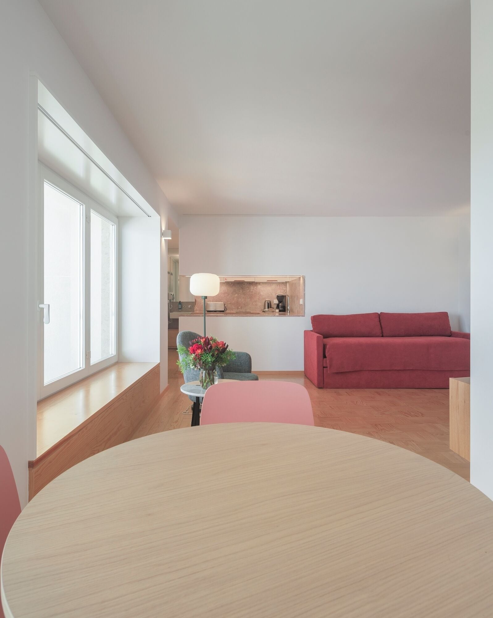 A modern living room with a dining table, a red sofa, a small kitchen area, and a large window.