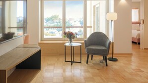 A modern living room with a wooden floor, a grey armchair, a small round table with a vase, and a large window with a city view.