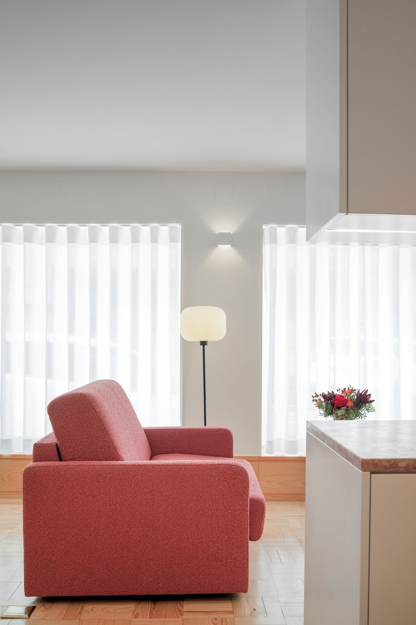 A modern living room with a red sofa, a floor lamp, and a kitchen counter with a flower arrangement.