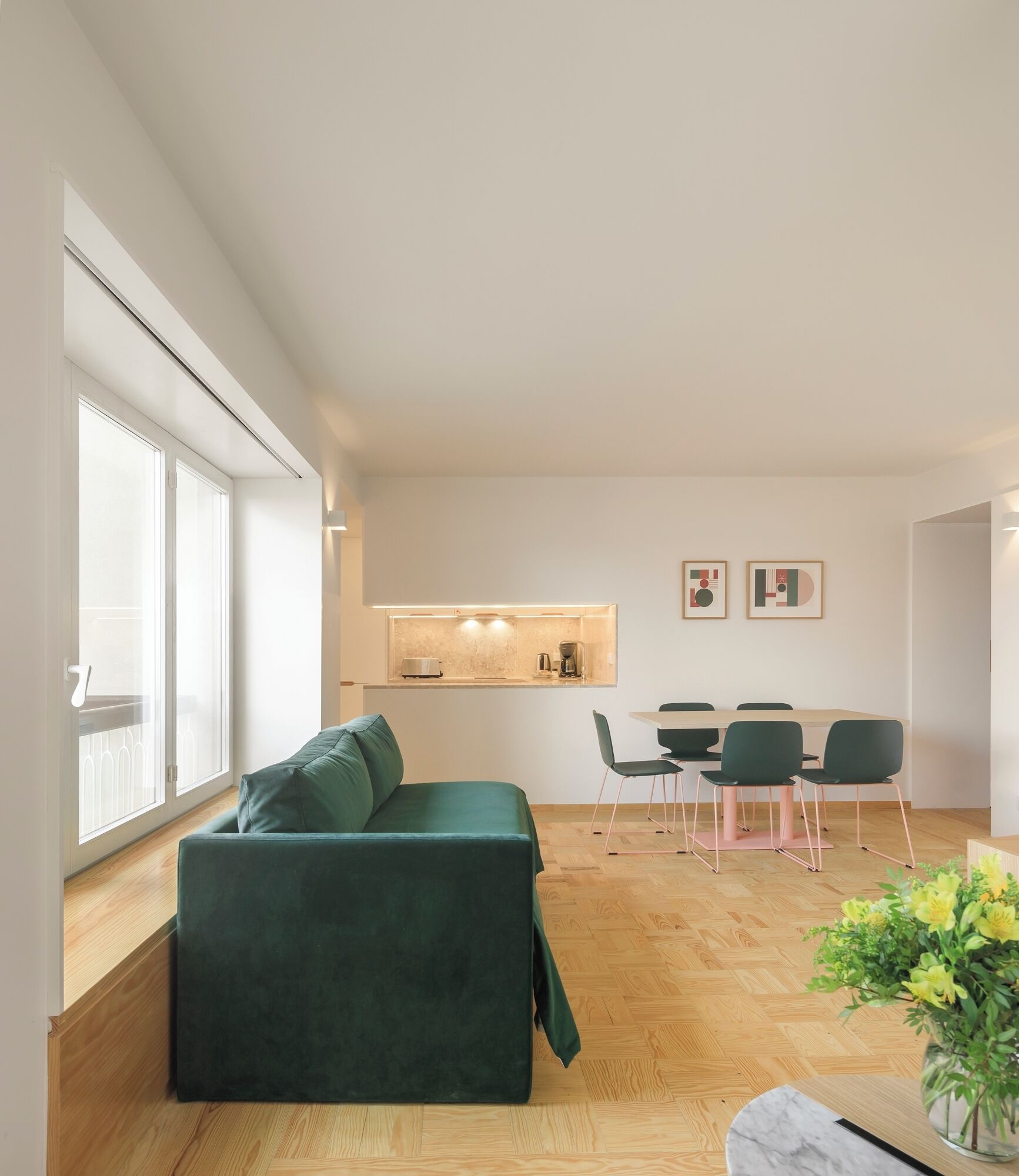 A modern living room with a green sofa, a dining table with chairs, and a kitchen area in the background.