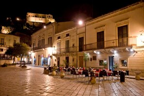 Un espace café en plein air avec des tables et des chaises, un bâtiment doté d’un balcon et, au fond, un bâtiment historique.