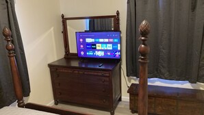 A bedroom with a wooden dresser, a bed with a patterned bedspread, a television on the dresser, and dark curtains.