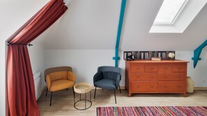 Une pièce avec un plafond en pente, une lucarne, une commode en bois, deux chaises, une petite table ronde et un tapis coloré.
