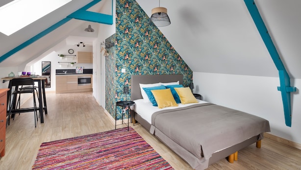 Une chambre avec un plafond en pente, un lit agrémenté d’oreillers bleus et jaunes, un tapis coloré et une lucarne.