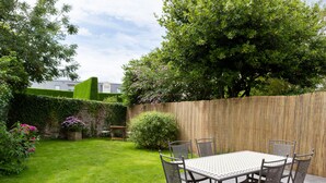 Une terrasse avec une table et des chaises, entourée d’une clôture en bois, d’une pelouse et de diverses plantes.