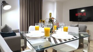 A modern living room with a glass dining table set for four, a sofa, a TV displaying "NETFLIX", and a bottle of champagne.