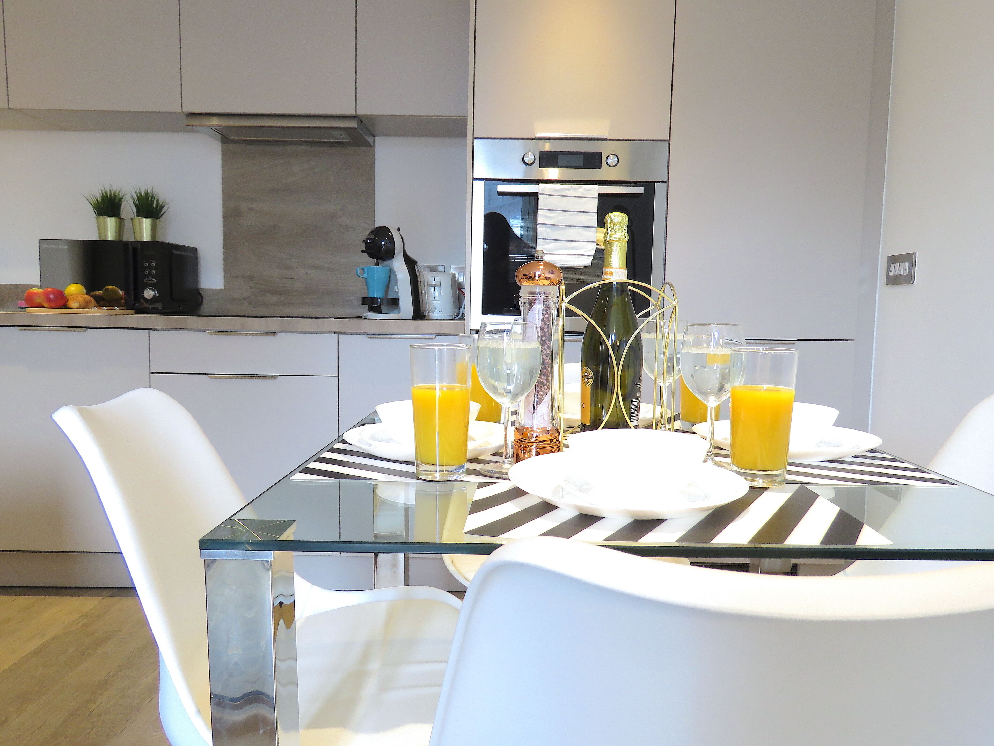 A modern kitchen with a glass dining table, white chairs, and a stainless steel oven.