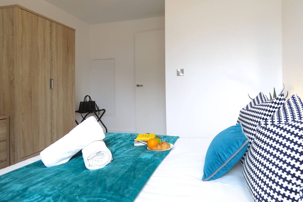 Een slaapkamer met een bed, een blauwgroene deken, een witte handdoek, een zwarte handtas, een boek en een bord met eten.