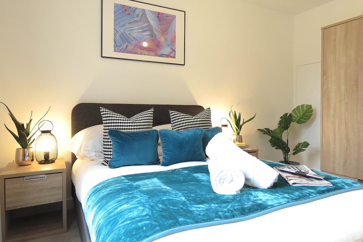 Een netjes opgemaakt bed met een blauwe deken, een hoofdbord en twee decoratieve kussens. Boven het bed hangt een ingelijste foto aan de muur.