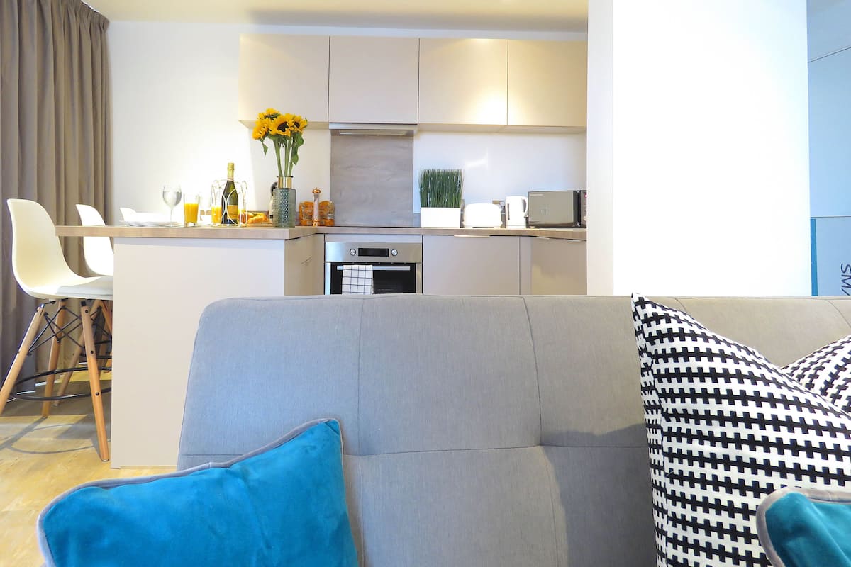Een moderne woonkamer met een grijze bank, een blauw kussen en een zwart-wit gestreept kussen. Op de achtergrond een keuken met een magnetron, een waterkoker en een vaas met gele bloemen.