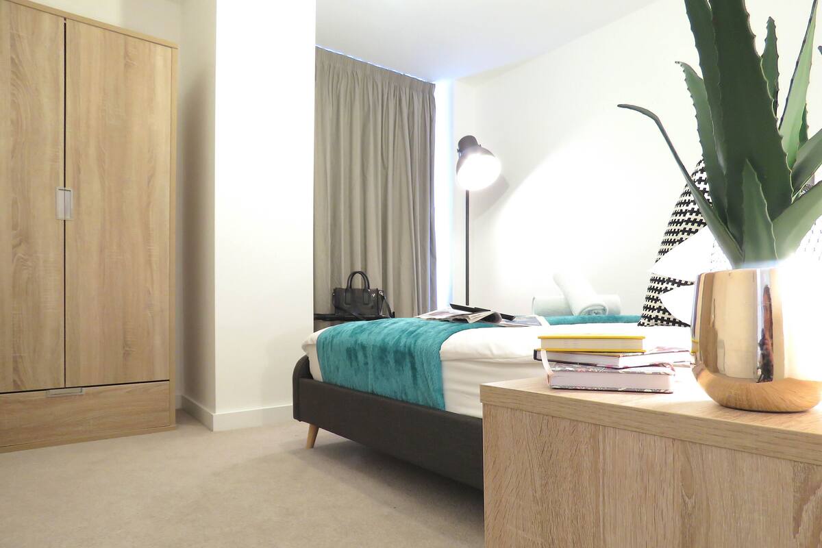 Een slaapkamer met een bed, een nachtkastje met boeken, een lamp, een kledingkast en een potplant.