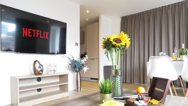 Een moderne woonkamer met een flatscreen televisie, een houten salontafel, boeken, een vaas met bloemen, en een eethoek op de achtergrond.