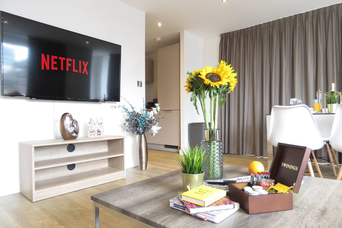 Een moderne woonkamer met een flatscreen televisie, een houten salontafel, boeken, een vaas met bloemen, en een eethoek op de achtergrond.