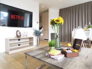 Un salon moderne avec un téléviseur à écran plat, une table basse en bois, des livres, un vase de fleurs et, au fond, un coin repas.