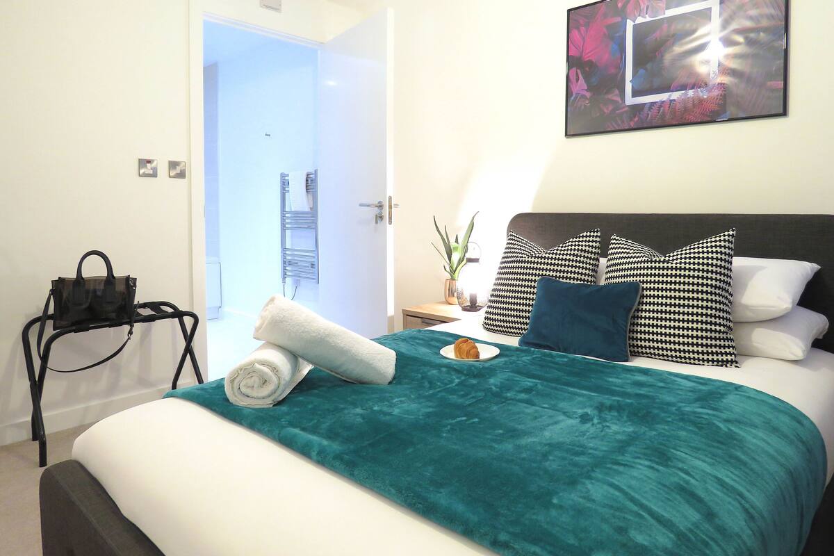 Een net opgemaakt bed met een blauwgroene spreidek, twee kussens en een opgerolde witte handdoek.