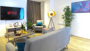 Een moderne woonkamer met een flatscreen waarop 'NETFLIX' staat, een bank, een salontafel met boeken en een plant, een vloerlamp en een groot abstract schilderij aan de muur.