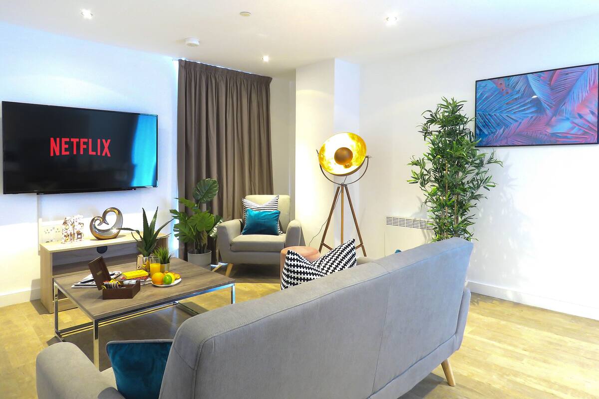 Een moderne woonkamer met een flatscreen waarop 'NETFLIX' staat, een bank, een salontafel met boeken en een plant, een vloerlamp en een groot abstract schilderij aan de muur.