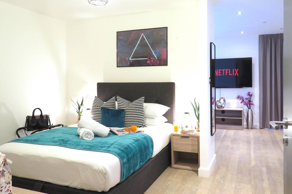 Een moderne hotelkamer met een groot bed, een nachtkastje met een lamp, een aan de muur bevestigde TV waarop 'NETFLIX' te zien is, en een schilderij boven het bed.