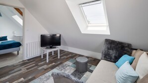 A modern loft bedroom with a sloped ceiling, a skylight, a television on a white side table, a patterned rug, and a sofa with cushions.