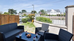 An outdoor patio with wicker furniture, a table with cups and a plate, and a view of a gated street with houses. - Le Repos- À 300 m de la plage, St Lunaire (Saint-Lunaire)