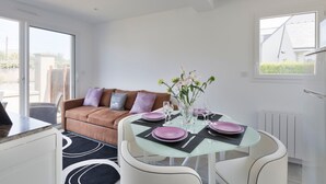 A modern dining area with a glass table set for four, a brown leather sofa with cushions, and a view of the outside through large windows. - Le Repos- À 300 m de la plage, St Lunaire (Saint-Lunaire)