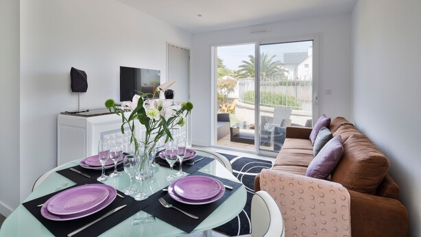 A modern dining area with a glass table set for four, a sofa, and a TV in the background. - Le Repos- À 300 m de la plage, St Lunaire (Saint-Lunaire)