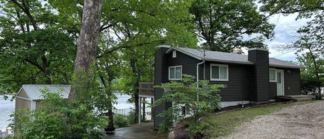 A dark-colored cabin with a deck, surrounded by trees and a body of water.