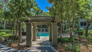Un sendero conduce a una zona de piscina enmarcada por una pérgola, rodeada de árboles y vegetación.