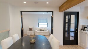 A dining area with a black table and white chairs, a small side table with a candle, and a view into a living room with a white sofa and a window. - Coastal modern home (Newport Beach)