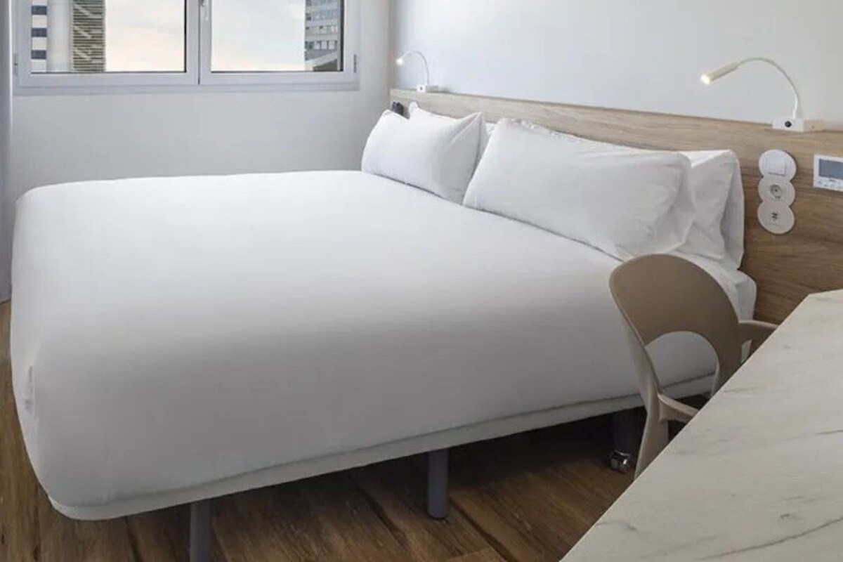 Una habitación de hotel moderna con una cama grande, piso de madera, una ventana con cortinas, una mesita de noche con lámpara y un panel de control en la pared.