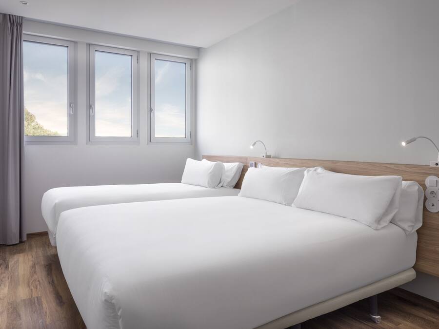 A modern hotel room with two single beds, a wooden headboard, bedside lamps, a phone, and a window with curtains.