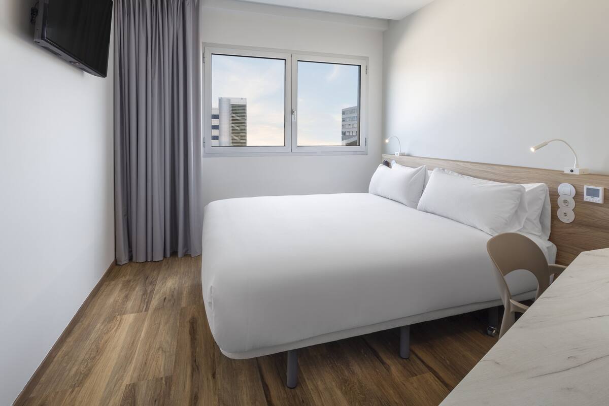 A modern hotel room with a large bed, a desk, a chair, a TV mounted on the wall, and a window with a view of buildings.