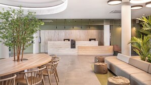 A modern interior with a wooden reception desk, a round dining table with chairs, a sofa, and a potted plant. - B&B Hotel Lisboa Aeroporto (Loures)