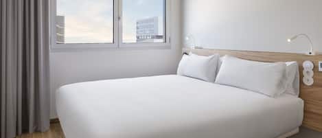 Una habitación de hotel moderna con una cama grande, cabecera de madera, mesita de noche con lámpara y ventana con cortinas.