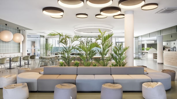 A modern lounge area with a large sectional sofa, round seating, and a central plant arrangement. - B&B Hotel Lisboa Aeroporto (Loures)