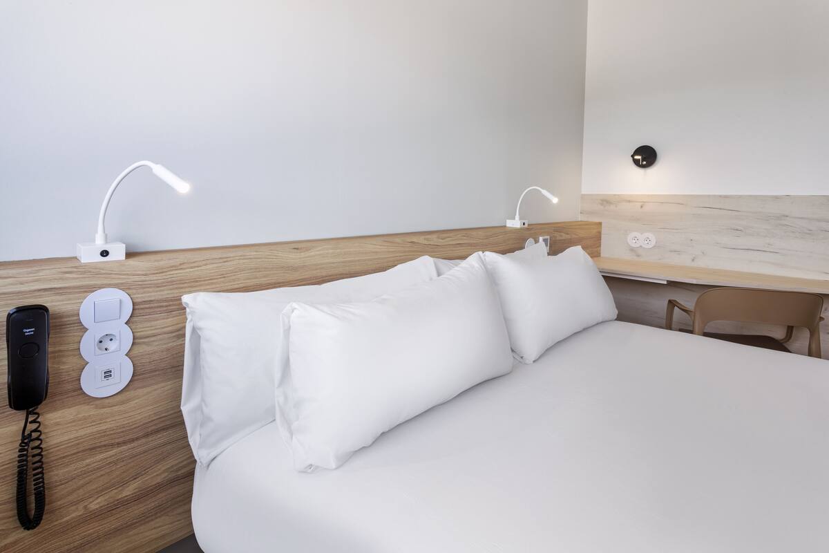 A hotel room with a wooden headboard, a bed with white bedding, a bedside table with a lamp, a phone, and a wall socket.