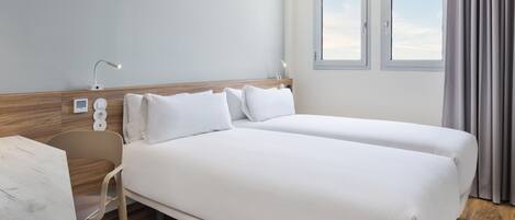 A hotel room with a double bed, a desk, a chair, a bedside table, a lamp, and a window with curtains.