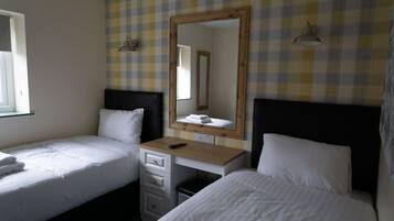 A hotel room with two single beds, a wooden desk with a mirror, a small drawer unit, and a window with curtains.