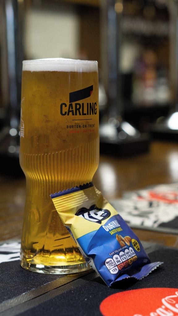 Ein Glas Bier mit dem Aufdruck 'CARLING BURTON-ON-TRENT' und einer Tüte Snacks daneben.