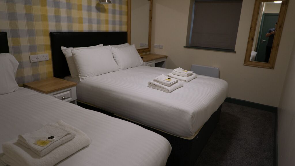 A hotel room with two beds, each with white linens and towels. There is a wooden nightstand with a lamp between the beds.