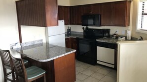 A compact kitchen with dark wooden cabinets, a granite countertop, a black microwave, a stove, and a refrigerator. - Villa with Great Ocean View (Atlantic Beach)