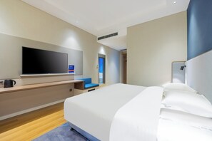 Una habitación de hotel moderna con una cama grande, un televisor de pantalla plana en la pared, un escritorio con una silla y una pequeña zona de estar con una silla azul.