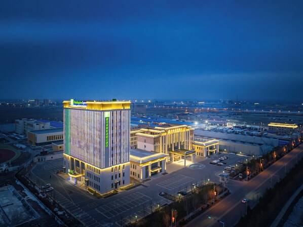 A modern hotel with a prominent vertical light strip design, a central courtyard, and a view of the surrounding cityscape at dusk. - Holiday Inn Express Horgos by IHG (Huocheng County)