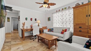 A living room with a wooden coffee table, a grey sofa, a wooden cabinet, a ceiling fan, and a desk with a lamp. - Davis Islands Urban Fiesta 2 Bedroom (Tampa)