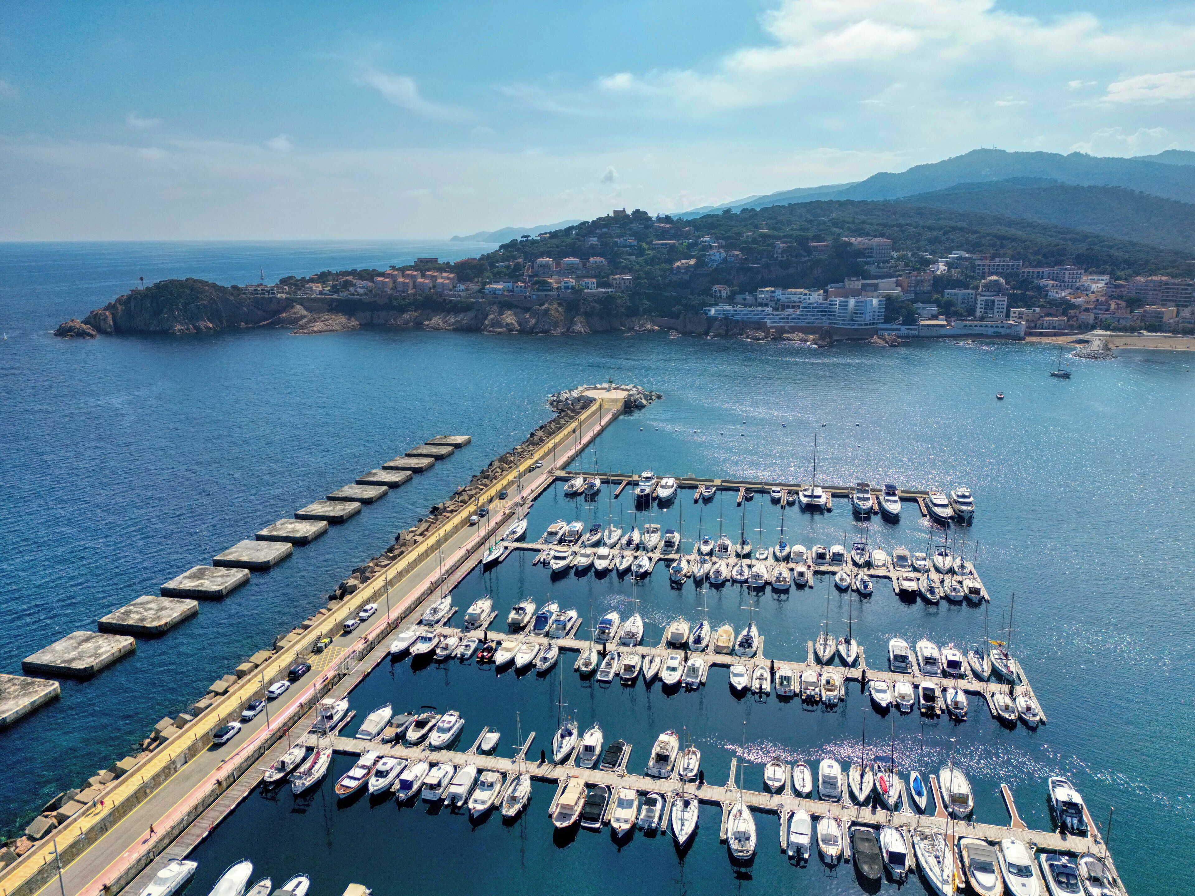 An aerial view of a marina with numerous boats docked, a long pier, and a mountainous backdrop.