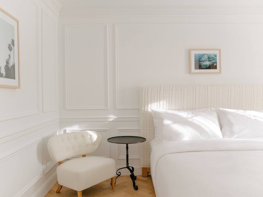 Une chambre d’hôtel avec un lit, une petite table ronde, un fauteuil blanc et un tableau encadré au mur.