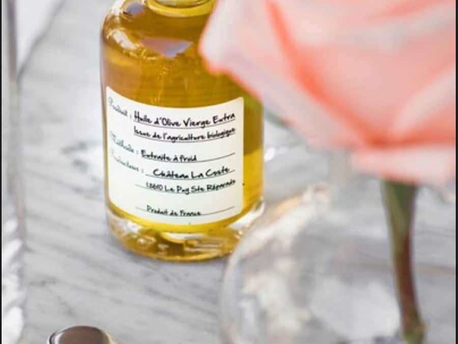 A bottle of olive oil with a label, a pink rose, and a glass on a marble surface.