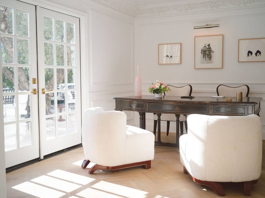 A room with two white upholstered chairs, a dark wooden desk with a vase of flowers, and framed artwork on the wall.