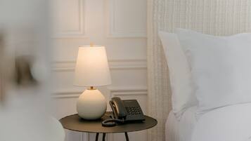 A neatly made bed with white linens, a bedside table with a lamp and a telephone, and a textured white wall in the background.